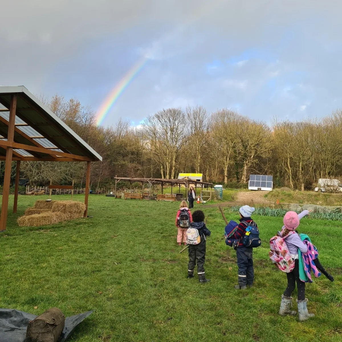 Four children pointing at a rainbow over the farm shelter and hay bales after a rain shower